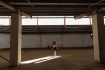 Vista frontal de una joven bailarina de ballet de raza mixta con zapatos puntiagudos de pie sobre sus dedos de los pies en el eje de la luz del sol con los brazos a sus lados mientras baila en una habitación vacía en un almacén abandonado - foto de stock