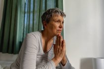 Вид збоку крупним планом зрілої кавказької жінки з коротким сивим волоссям сидить на ліжку вдома руки разом, як якби в молитві, дивлячись — стокове фото
