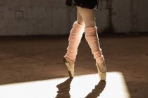 Unterteil einer Balletttänzerin, die im Sonnenlicht in einem leeren Raum einer verlassenen Lagerhalle in Beinstulpen und Spitzenschuhen auf den Zehen steht — Stockfoto