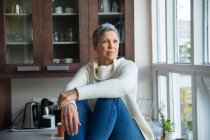 Front view close up of a mature Caucasian woman with short grey hair sitting on the counter in her kitchen looking out of the window — Stock Photo