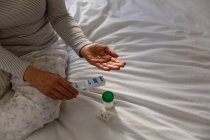 Вид спереду середня частина жінки сидить на її ліжку вдома, тримаючи дві таблетки і щотижневу коробку для таблеток, з іншими контейнерами ліків на ліжку поруч з нею — стокове фото