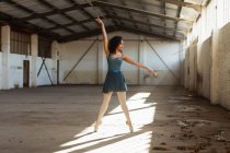Vue de face d'une jeune danseuse de ballet mixte debout sur ses orteils les bras levés dans un rayon de soleil en dansant dans une pièce vide dans un entrepôt abandonné — Photo de stock
