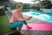 Vue latérale d'une femme blanche mature aux cheveux gris court portant des vêtements de sport à l'aide d'un smartphone assis près de la piscine dans son jardin après une séance de yoga, avec une vue rurale en arrière-plan — Photo de stock