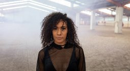 Portrait of a young mixed race woman with shoulder length curly hair wearing a black mesh top looking straight to camera in an abandoned warehouse — Stock Photo