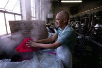 Side view of a senior mixed race man wearing an apron holding and shaping the top of a hat that has been steamed in the workshop at a hat factory, with equipment visible in the background — Stock Photo