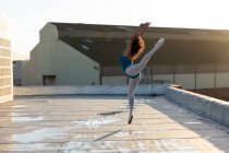 Vista lateral de una joven bailarina de ballet de raza mixta saltando con los brazos y una pierna levantada, en la azotea de un edificio urbano, retroiluminada por la luz del sol - foto de stock