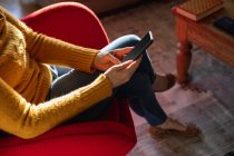 Hochstehender Unterteil einer Frau, die in einem roten Sessel in ihrem Wohnzimmer sitzt und ihr Smartphone benutzt, mit überkreuzten Beinen — Stockfoto