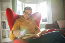 Vue de face gros plan d'une femme blanche mature aux cheveux gris court portant des lunettes assise dans un fauteuil rouge dans son salon lisant un livre, rétro-éclairée par la lumière du soleil depuis la fenêtre — Photo de stock