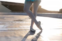 Нижняя часть балетной танцовщицы в колготках и пуантах, стоящая со скрещенными ногами на крыше городского здания, освещенная солнечным светом — стоковое фото