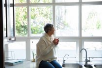Vista lateral de una mujer caucásica madura con el pelo gris corto sentado en el mostrador en su cocina sosteniendo una taza de café y mirando por la ventana, hay árboles afuera y el sol está brillando - foto de stock