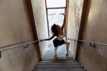 Vista lateral elevada de una joven bailarina de ballet de raza mixta bailando en una escalera en un almacén abandonado - foto de stock