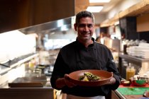 Retrato de um cozinheiro masculino caucasiano de meia idade sorridente segurando um prato de comida preparada em uma cozinha de restaurante — Fotografia de Stock