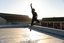 Vista lateral de una joven bailarina de ballet de raza mixta saltando en el aire desde una pierna en una pose de ballet con un brazo en el aire, en la azotea de un edificio urbano, retroiluminada por la luz del sol - foto de stock