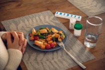 Gros plan des mains de la femme reposant sur la table, avec de la nourriture sur une assiette, des bouteilles de comprimés et de pilules et un verre d'eau devant elle — Photo de stock