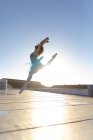 Вид сбоку на молодую балетную танцовщицу смешанной расы в синей пачке и пуантах, прыгающую с поднятой рукой, на крыше городского здания, освещенного солнечным светом — стоковое фото