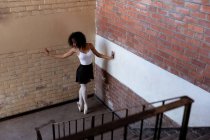 Vue latérale surélevée d'une jeune danseuse de ballet mixte tenant une pose de danse debout sur ses orteils dans un coin sur un escalier atterrissant dans un entrepôt abandonné — Photo de stock