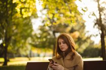 Frau benutzt Handy, während sie auf einer Bank im Park sitzt. — Stockfoto