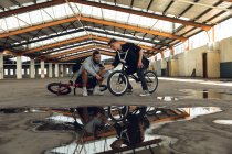 Vue de face de deux jeunes hommes blancs adultes assis sur des vélos BMX parlant, l'un montrant l'autre son smartphone dans un entrepôt abandonné — Photo de stock