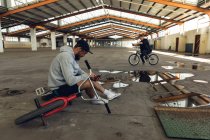 Vista lateral de dois jovens adultos caucasianos sentados em bicicletas BMX usando smartphones em um armazém abandonado — Fotografia de Stock