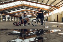 Frontansicht von zwei jungen erwachsenen kaukasischen Männern, die auf BMX-Fahrrädern sitzen, miteinander reden und Smartphones in einer verlassenen Lagerhalle benutzen — Stockfoto