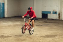 Вид спереди молодого кавказца, катающегося на заднем колесе велосипеда BMX, практикующего трюки на заброшенном складе — стоковое фото