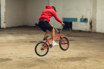 Seitenansicht eines jungen kaukasischen Mannes, der einen Hasensprung auf einem BMX-Fahrrad macht, während er in einer verlassenen Lagerhalle Tricks übt — Stockfoto