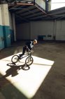 Vista lateral de cerca de un joven hombre caucásico deslizándose hacia los lados para detenerse en un pozo de luz solar en una bicicleta BMX mientras practica trucos en un almacén abandonado - foto de stock