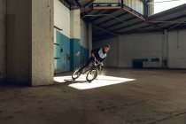Vista lateral de um jovem caucasiano deslizando de lado para uma parada em uma bicicleta BMX em um eixo de luz solar, enquanto pratica truques em um armazém abandonado — Fotografia de Stock