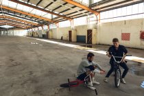Vue de face de deux jeunes hommes caucasiens adultes assis sur des vélos BMX se parlant et utilisant des smartphones dans un entrepôt abandonné — Photo de stock