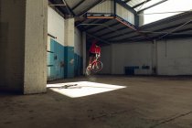 Seitenansicht eines jungen kaukasischen Mannes, der einen Sprung auf einem BMX-Fahrrad macht und in einer verlassenen Lagerhalle den Lenker dreht — Stockfoto