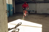 Vista frontal de un joven caucásico haciendo un salto en una bicicleta BMX en un almacén abandonado - foto de stock
