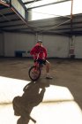 Portrait d'un jeune homme caucasien barbu portant une casquette de baseball, un short et un haut rouge assis sur un vélo BMX regardant la caméra dans un entrepôt abandonné, la lumière du soleil projetant son ombre devant lui — Photo de stock