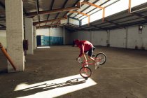 Vue latérale d'un jeune homme caucasien se balançant sur la roue avant d'un vélo BMX dans un rayon de soleil tout en pratiquant des tours dans un entrepôt abandonné — Photo de stock