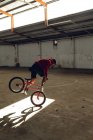 Side view of a young Caucasian man balancing on the front wheel of a BMX bike in a shaft of sunlight while practicing tricks in an abandoned warehouse — Stock Photo