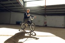 Seitenansicht eines jungen kaukasischen Mannes, der auf einem BMX-Fahrrad in einer verlassenen Lagerhalle sitzt und neben sich das Sonnenlicht wirft — Stockfoto