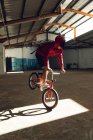 Nahaufnahme eines jungen kaukasischen Mannes, der auf dem Vorderrad eines BMX-Fahrrads in einem Sonnenlicht balanciert, während er in einer verlassenen Lagerhalle Tricks übt — Stockfoto