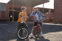 Vista frontal de cerca de dos jóvenes caucásicos sentados en bicicletas BMX hablando, uno mostrando al otro su teléfono inteligente fuera de un almacén abandonado, retroiluminado por la luz del sol - foto de stock