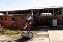 Side view of a young Caucasian man wearing sunglasses balancing on a barrel on the front wheel of a BMX bike outside an abandoned warehouse in the sun — Stock Photo