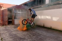 Vista lateral de um jovem caucasiano usando óculos de sol pulando sobre granadas de fumaça verde e laranja de pé em uma bicicleta BMX fora de um armazém abandonado ao sol — Fotografia de Stock