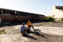 Rück- und Vorderansicht von zwei jungen kaukasischen Männern mit Sonnenbrille, die mit BMX-Fahrrädern vor einer verlassenen Lagerhalle in der Sonne sitzen und reden — Stockfoto