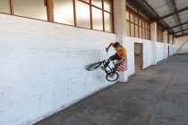 Vista frontal de um jovem caucasiano que caminha em uma bicicleta BMX em um armazém abandonado — Fotografia de Stock