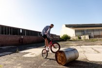 Side view of a young Caucasian man wearing sunglasses touching a barrel with the front wheel of a BMX bike outside an abandoned warehouse in the sun — Stock Photo