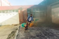 Frontansicht eines jungen kaukasischen Mannes mit Sonnenbrille, der ein BMX-Fahrrad mit grünen und orangefarbenen Rauchgranaten vor einer verlassenen Lagerhalle in der Sonne fährt — Stockfoto