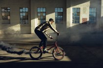 Side view of a young Caucasian man riding a BMX bike with a grey smoke grenade attached to it, in an abandoned warehouse — Stock Photo