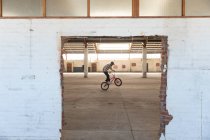Side view of a young Caucasian man balancing on the back wheel of a BMX bike while practicing tricks in an abandoned warehouse, seen through a broken doorway — Stock Photo