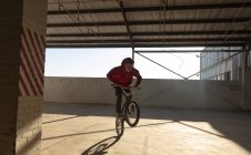 Vorderansicht eines jungen kaukasischen Mannes, der ein BMX-Fahrrad fährt und auf dem Hinterrad balanciert, während er in einer verlassenen Lagerhalle Tricks übt — Stockfoto