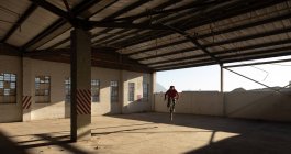 Vue de face d'un jeune homme caucasien en vélo BMX et sautant tout en pratiquant des tours dans un entrepôt abandonné — Photo de stock