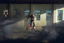Vista lateral de un joven caucásico saltando en una bicicleta BMX con una granada de humo gris, en un almacén abandonado - foto de stock