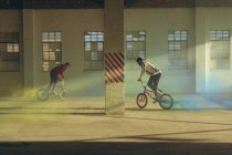 Side view of two young Caucasian men riding around through shafts of sunlight on BMX bikes with yellow and blue smoke grenades attached to them, in an abandoned warehouse — Stock Photo