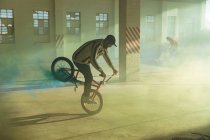 Side view of two young Caucasian men riding and doing tricks on BMX bikes with yellow and blue smoke grenades attached to them, in an abandoned warehouse — Stock Photo